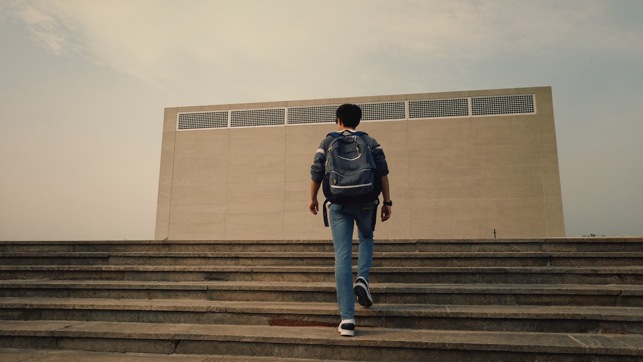A student with a backpack walking up wide stone steps toward a building in early morning light