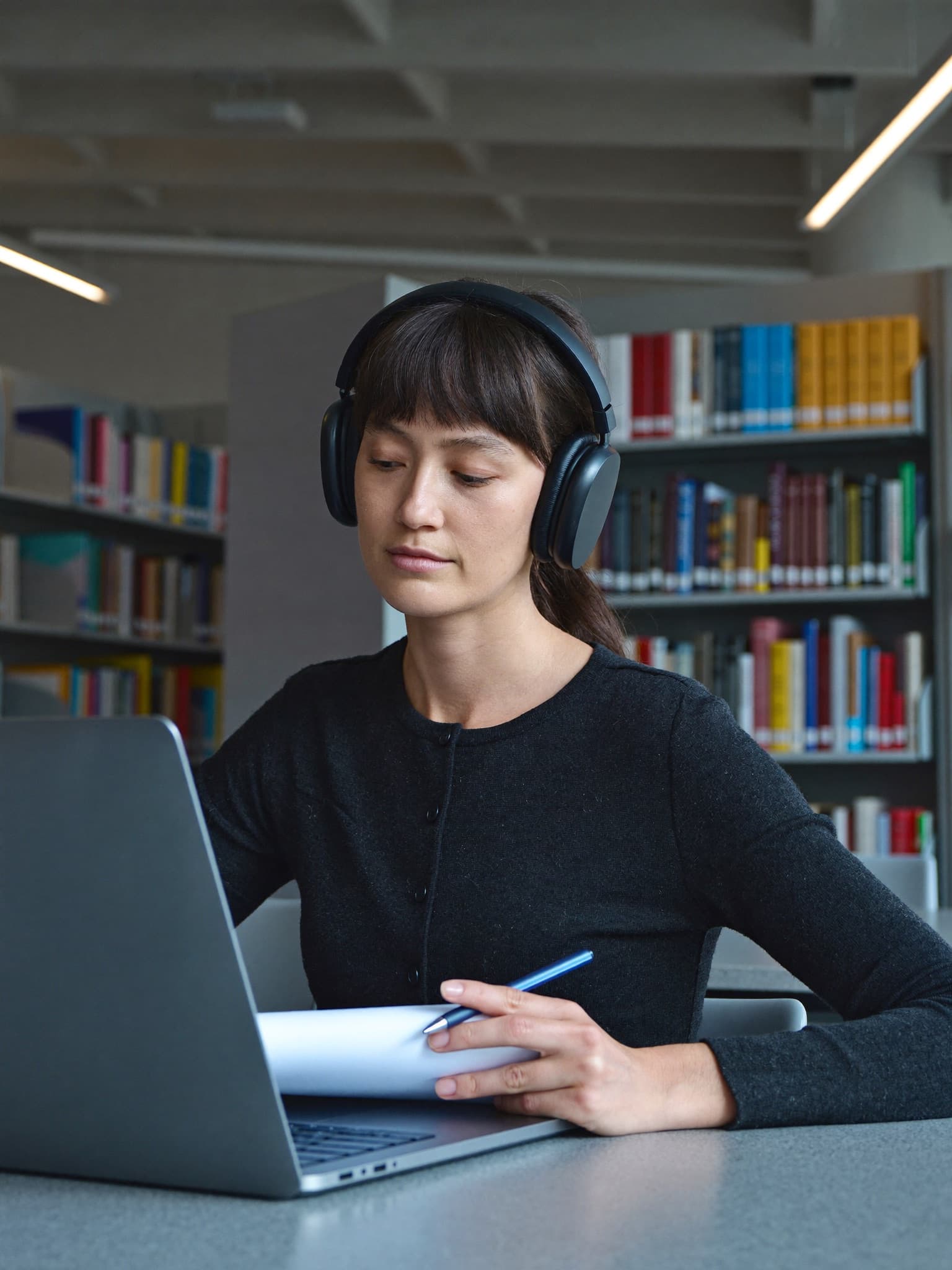 A learner wearing over-ear headphones taking notes beside a laptop in a modern university library